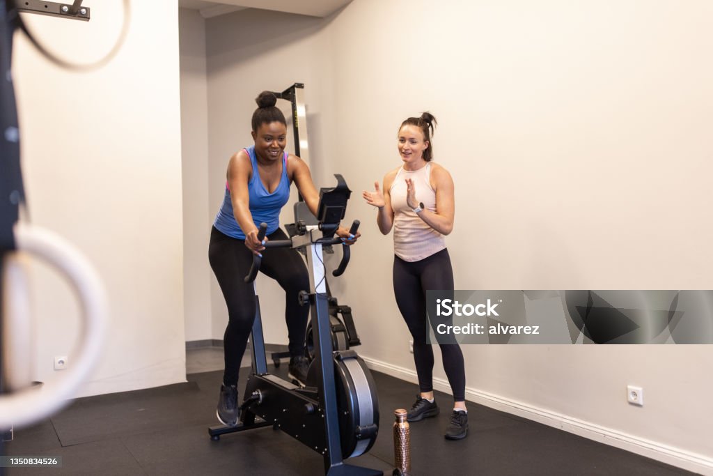 Personal trainer motivating woman exercising on a stationary bike at the fitness studio Female trainer clapping and motivating a woman exercising on a stationary bicycle at a fitness studio. Woman doing cardio workout at gym with instructor. Active Lifestyle Stock Photo