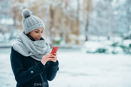 Beautiful young woman taking selfie with a smartphone and wearing scarf and a a hat on a cold winter day during snow