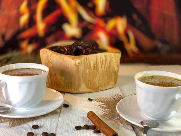 On the wooden table by the fireplace are white cups with coffee and gingerbread cookies in a wooden bowl