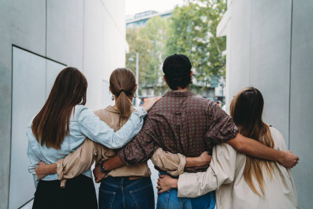 Rear view of four friends embracing together Rear view of four friends embracing together. They are standing. Unity concept. arm in arm stock pictures, royalty-free photos & images