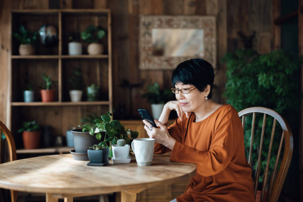 テーブルに座って笑顔のアジア人女性、ネットサーフィン、自宅でスマートフォンでオンラインショッピング。高齢者と技術 - coffee plant ストックフォトと画像