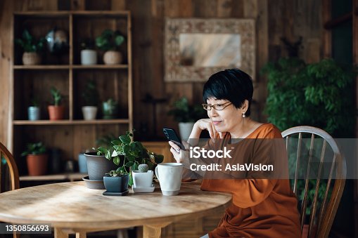 istock Smiling senior Asian woman sitting at the table, surfing on the net and shopping online on smartphone at home. Elderly and technology 1350856693