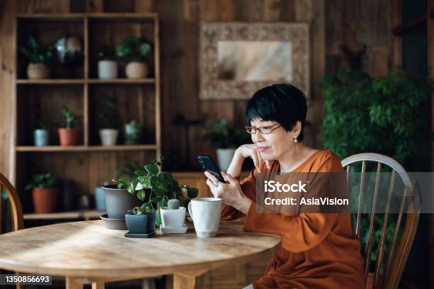 Smiling Senior Asian Woman Sitting At The Table Surfing On The Net And Shopping Online On Smartphone At Home Elderly And Technology Stock Photo - Download Image Now