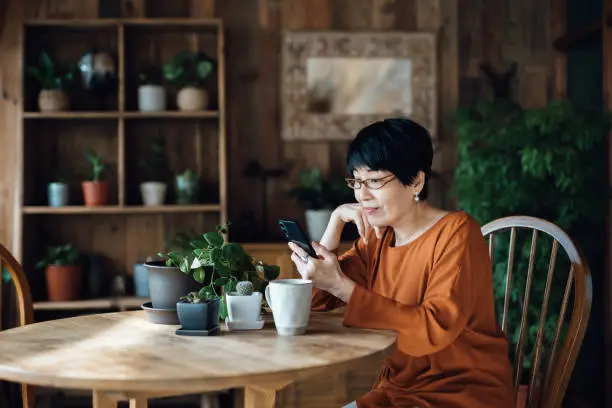 Photo of Smiling senior Asian woman sitting at the table, surfing on the net and shopping online on smartphone at home. Elderly and technology