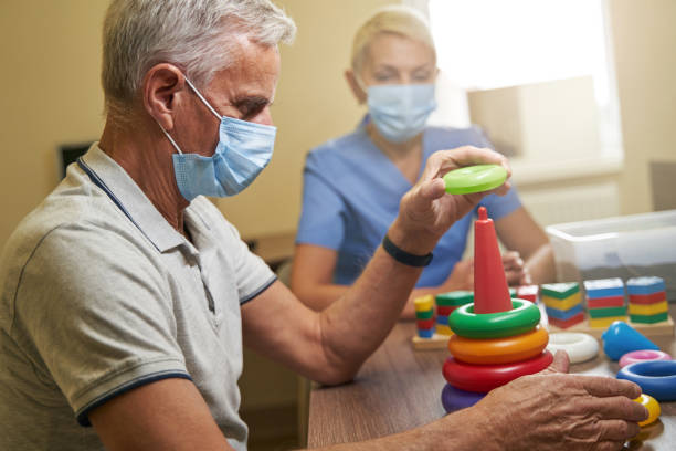 Shot of mature person while recovery of motor skills indoors Stroke patient and professional therapist both wearing protective masks while doing ergotherapy with games to stimulate memory in rehabilitation room motor living stock pictures, royalty-free photos & images