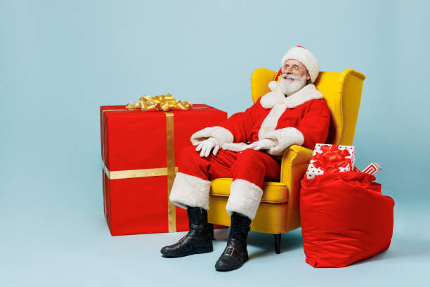 Full body old Santa Claus man 50s wear Christmas hat red suit sit in armchair near gifts bag isolated on plain blue background studio. Happy New Year 2022 celebration merry ho x-mas holiday concept el anciano de cuerpo completo de santa claus de los años 50 usa sombrero de navidad traje rojo sentado en el sillón cerca de la bolsa de regalos aislada en el estudio de fondo azul liso. feliz año nuevo 2022 celebración feliz ho concepto de vacaciones
