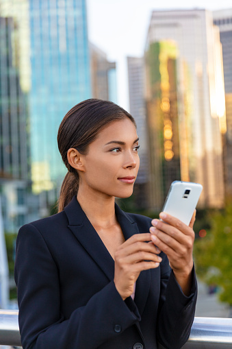 Asian business woman texting on mobile phone looking away to the side city background. Office building outdoors working people. Chinese businesswoman using smartphone with hand.