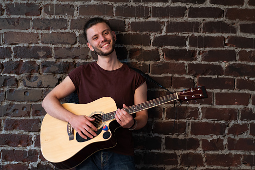Man with acoustic guitar against brick wall playing music singing songs enjoy life Handsome caucasian male guitar player practice play musical instrument home loft interior Creative lifestyle hobby
