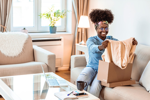 Young curly hair satisfied happy african woman shopaholic customer sitting on sofa unpack parcel.