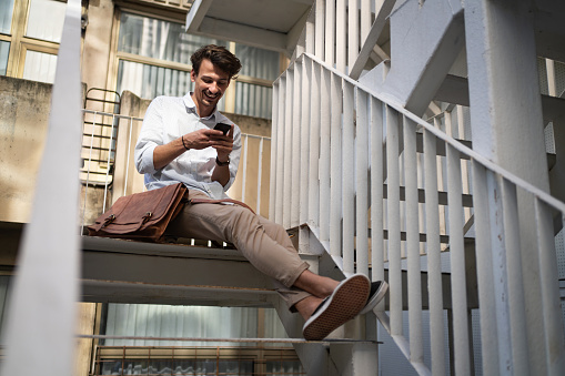 Carefree and modern young Caucasian businessman sitting on the stairs and surfing the net or text messaging on his mobile phone