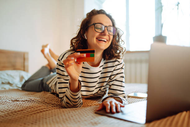 compras en línea en casa. una joven sostiene una tarjeta de crédito y usa una computadora portátil. - black friday fotografías e imágenes de stock