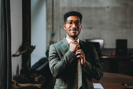 Portrait of a young adult businessman in his office. He's standing in a modern industrial loft. He's tying his tie.