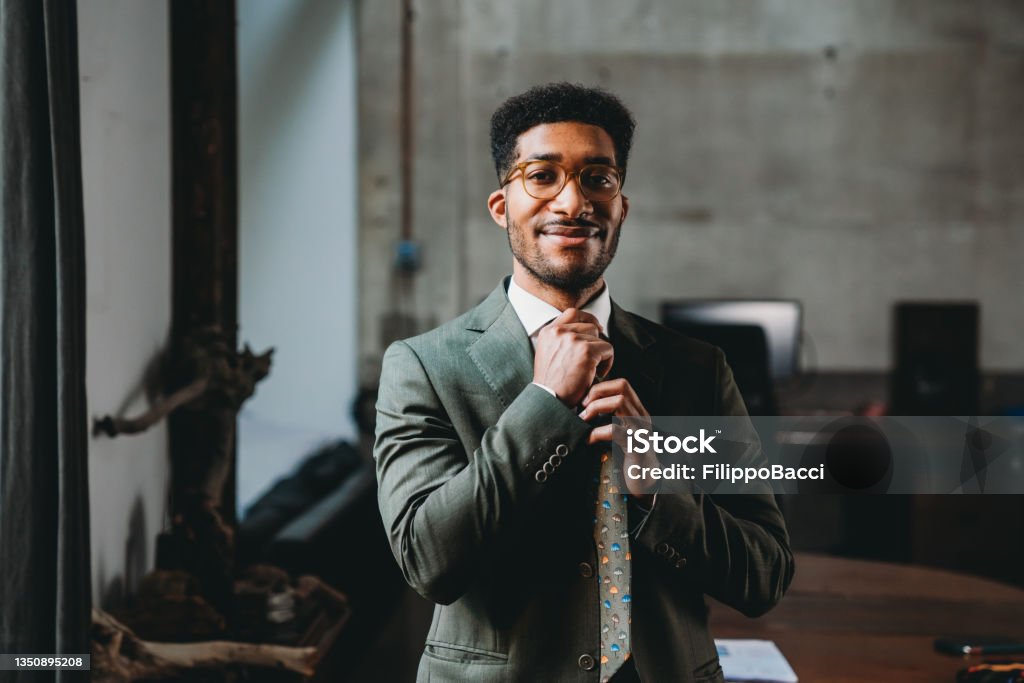 Portrait of a young adult businessman in his office Portrait of a young adult businessman in his office. He's standing in a modern industrial loft. He's tying his tie. Necktie Stock Photo