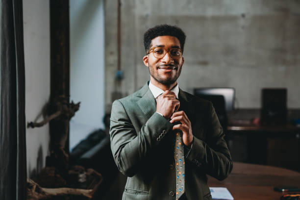 Portrait of a young adult businessman in his office Portrait of a young adult businessman in his office. He's standing in a modern industrial loft. He's tying his tie. man adjusting tie stock pictures, royalty-free photos & images
