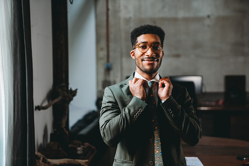 Portrait of a young adult businessman in his office. He's standing in a modern industrial loft. He's tying his tie.