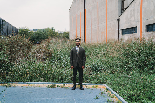 A young adult businessman is standing in a public park in the city. Industrial area. Uncultivated lawn in the background.