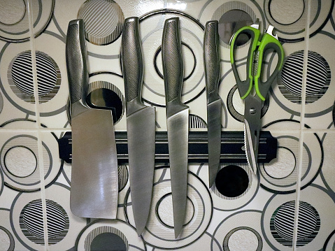 Magnetic knife holder. A set of steel knives. Kitchen scissors. Metallic kitchen tiles in the interior.