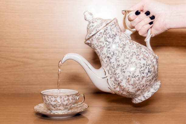 Tea set with golden ornaments. Hand holding a teapot pouring hot water in a small teacup. Wooden background