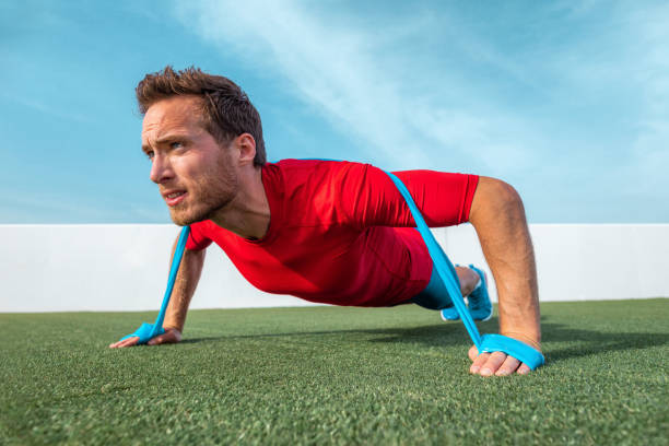 Resistance bands fit man doing pushups with elastic band as extra difficulty at outdoor gym park. Athlete doing dynamic workout pulling rubber elastics plyometrics exercises. las bandas de resistencia se ajustan al hombre haciendo flexiones con banda elástica como dificultad adicional en el parque del gimnasio al aire libre. atleta haciendo entrenamiento dinámico tirando de elásticos de goma ejercicios pliométricos