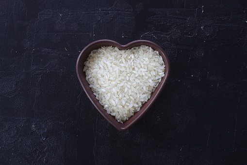 Isolated handful of raw white rice in the wood bowl in the shape of heart on black stone background