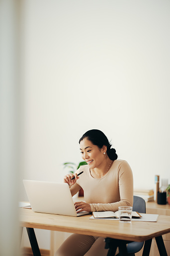 Beautiful smiling Asian businesswoman reading business report on a laptop computer while sitting at desk in a living room