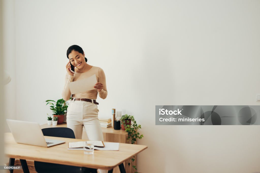 Cheerful Business Woman Analyzing Document and Talking on Mobile Phone Successful Asian businesswoman holding business report on a paper and speaking on the phone while standing in the modern office Using Phone Stock Photo
