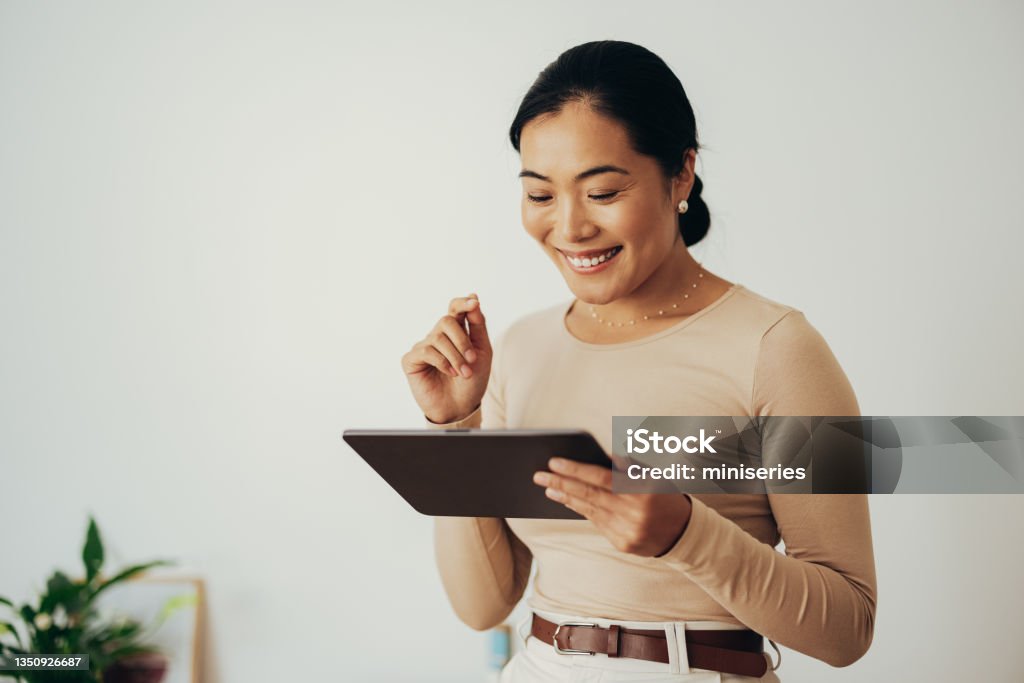 Happy Business Woman Using Digital Tablet at Home Beautiful smiling Asian businesswoman reading something on a digital tablet while standing in a modern office Digital Tablet Stock Photo