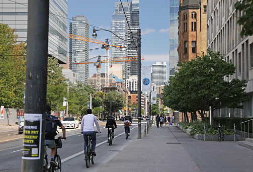 Toronto, Canada - September 3, 2021: Cyclists travel east on a bicycle lane on College Street, a major arterial route through downtown. University of Toronto education buildings line the 100-block College Street. Colorful street banners show the University's proud research history.  \n\nConstruction cranes stand near Queens Park in the background. Summer afternoon.