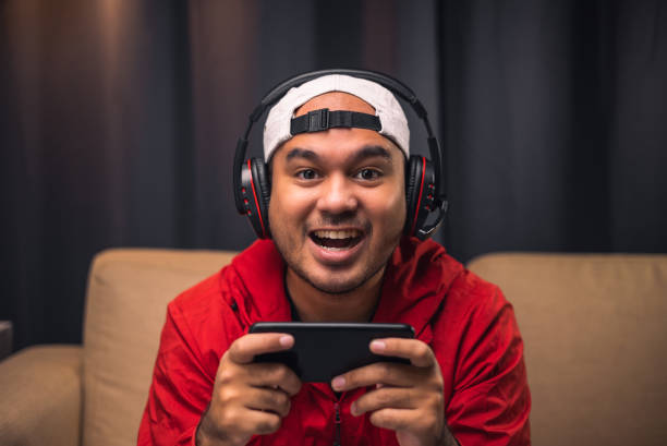 istock Playing video game on smartphone. Young asian handsome man sitting on sofa holding cellphone in living room. Happiness Streamer Indian man wearing headset playing game online in the darkroom. 1350963192