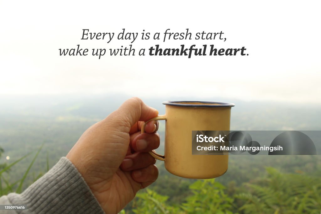 Person holding yellow cup of tea or coffee in hand against mountain view with a morning thankful inspirational message. Grateful inspirational quote - Every day is a fresh start, wake up with a thank heart. Hand holding tea or coffee cup on foggy mountain background in a misty morning. Morning relax with tea outdoor. Inspirational Quote Stock Photo