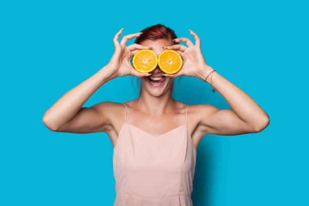 Lovely red haired woman is covering her eye with sliced lemons posing on a blue studio wall Lovely red haired woman is covering her eye with sliced lemons posing on a blue studio wall woman applying wallcovering stock pictures, royalty-free photos & images