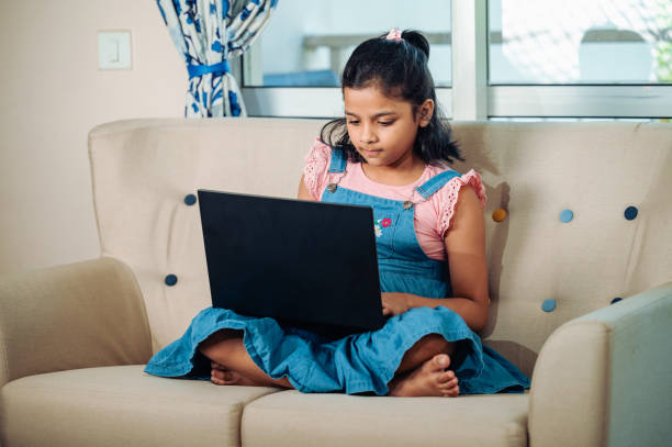 retrato de menina sorridente usando laptop enquanto senta no sofá em casa. - indian girls audio - fotografias e filmes do acervo
