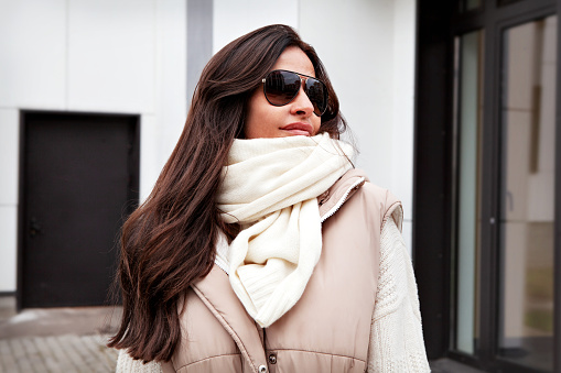 Close up Portrait of beautiful woman looking away on street. Stylish Model with long brunette hair wearing sunglasses, knitted white scarf and sweater, beige vest