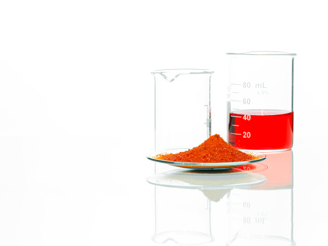 Potassium Ferricyanide in Chemical Watch Glass place next to red liquid chemical in beaker on white laboratory table. Side View