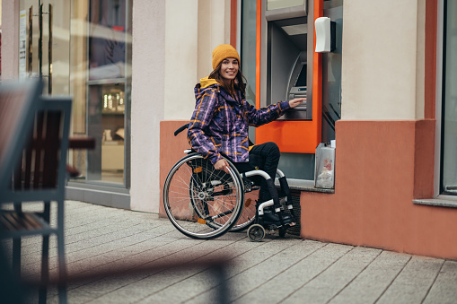 Young beautiful woman in wheelchair using credit card and atm machine while withdrawing money