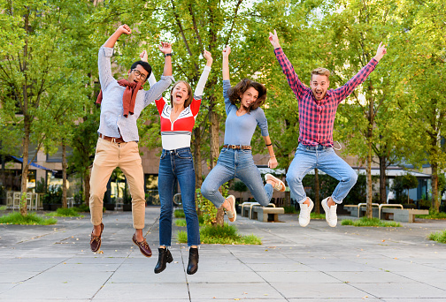 Group of exuberant diverse young friends cheering and leaping in the air with arms raised in celebration outdoors in a park