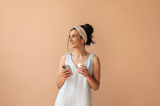 Beautiful female person taking break during studio reconstruction, drinking coffee, holding her smartphone and looking away