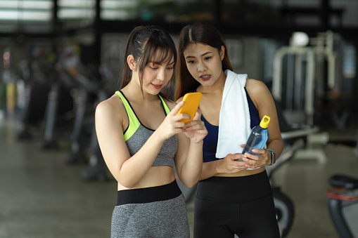 Two female best friends at the gym, looking at smartphone screen, using phone to check her social media and take some rest after workout.