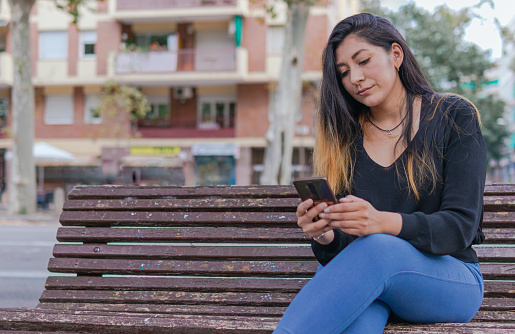 A latin woman is sitting with crossed legs on a bench of an avenue with a neutral expression using her phone