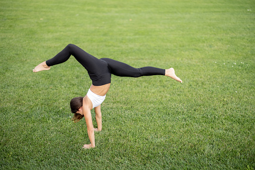 Young girl practicing yoga on green grass of meadow at sunny day. Concept of healthy lifestyle. Idea of relaxation. Side view of woman barefoot and wearing sportswear