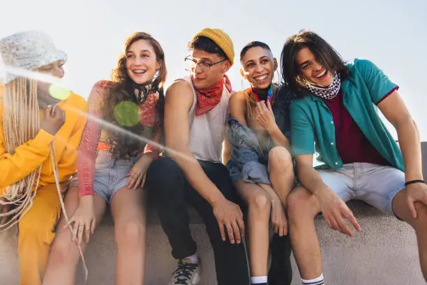 Hanging out in the sun. Group of happy friends laughing cheerfully while sitting together on a wall in the city. Multiethnic youngsters having fun together during the coronavirus pandemic.