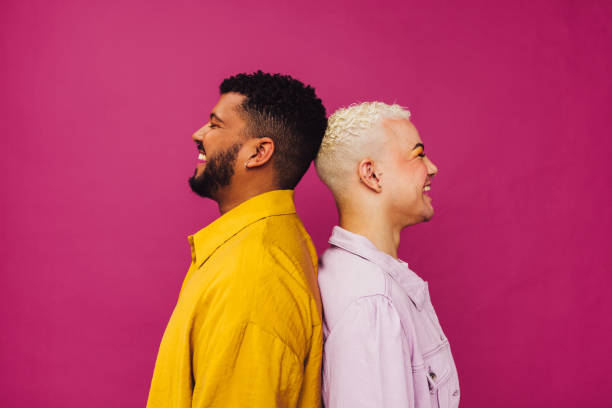 Cheerful gay couple standing together in a studio Cheerful gay couple standing together in a studio. Sideview of a beautiful gay couple with piercings standing back to back against a purple background. Young LGBTQ+ couple smiling happily. gay man stock pictures, royalty-free photos & images