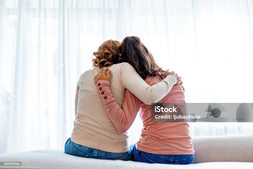 Rear view of mother and daughter embracing sitting on bed Rear back view of a mother and daughter embrace sitting on bed at home, older sister consoling younger teen, girl suffers from unrequited love share secrets trustworthy person relative people concept Mother Stock Photo