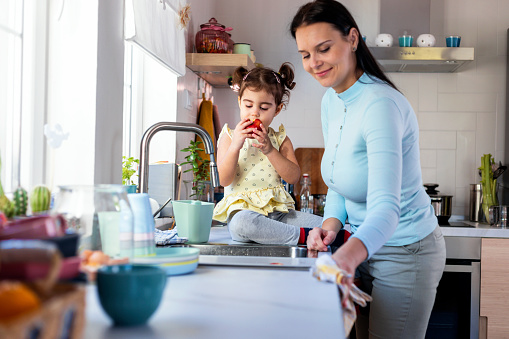 Beautiful woman with her cute little daughter playing in kitchen. They are eating apples, cleaning  and spending quality time together in domestic kitchen