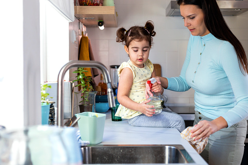 Beautiful woman with her cute little daughter playing in kitchen. They are cleaning kitchen counter  and having lot of fun. Family  spending quality time together in domestic kitchen.