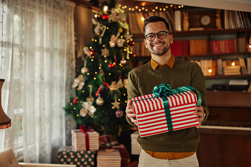 Smiling young adult man with eyeglasses standing nearby Christmas tree in the living room and holding wrapped present