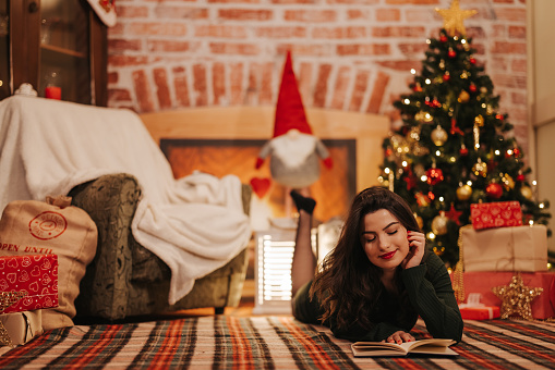 Beautiful young woman lying on floor and reading book during Christmas and New Year holidays at home