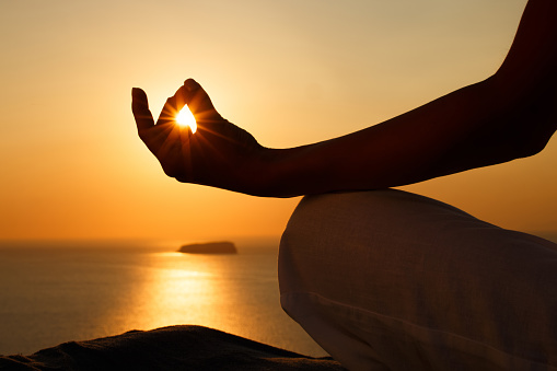 Close up of unrecognizable person doing Yoga relaxation exercises in Lotus position on a hill above the sea at sunset. Copy space.