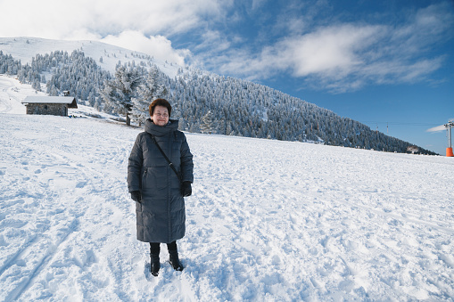 Senior beautiful smiling woman in long grey coat at snowy slope enjoying winter holidays at ski resort with splendid snow-covered mountains view in background. Winter vacation