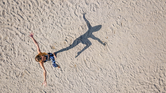 Drone view on woman practicing yoga on the sand, standing on one leg with the other up bent in knee, assuming One legged mountain pose, casting shadow in shape of her figure, practicing yoga outdoors